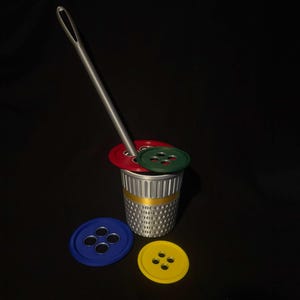 Op de afbeelding: Een zilveren vingerhoed met een gouden band en een grote naald, omringd door kleurrijke knopen. De knopen zijn rood, groen, blauw en geel. De naald is in de vingerhoed gestoken, wat een naaithema suggereert.