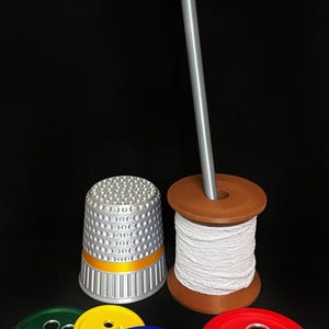 Op de afbeelding: Een verzameling naaigerei en accessoires, waaronder een zilveren vingerhoed met een gele band, een bruine spoel met wit garen, een grote naald en kleurrijke knopvormige onderzetters in groen, geel, blauw en rood.