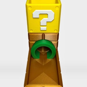 Op de afbeelding: Een gele, kubusvormige houder met een wit vraagteken aan de voorzijde, bevestigd op een metallic bronskleurige basis met een centrale groene cirkelvormige boog. De structuur heeft een holle opening aan de bovenzijde en een plat platform.