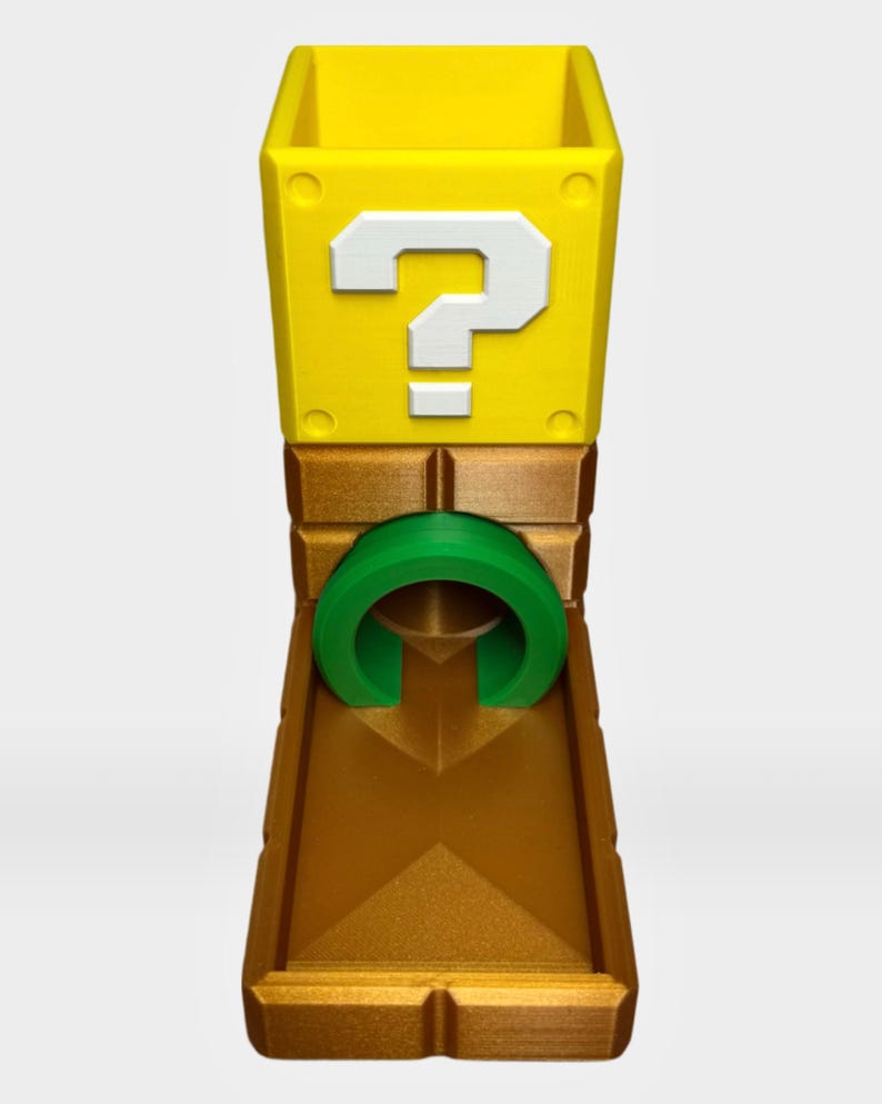 Op de afbeelding: Een gele, kubusvormige houder met een wit vraagteken aan de voorzijde, bevestigd op een metallic bronskleurige basis met een centrale groene cirkelvormige boog. De structuur heeft een holle opening aan de bovenzijde en een plat platform.