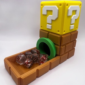 Op de afbeelding: Een 3D-geprinte dobbelsteentoren met bak, bestaande uit een geel vraagtekenblok op een bruine bakstenen basis met een groene buisopening. Verschillende doorschijnende rood-witte veelzijdige dobbelstenen met gouden cijfers liggen in de bak.