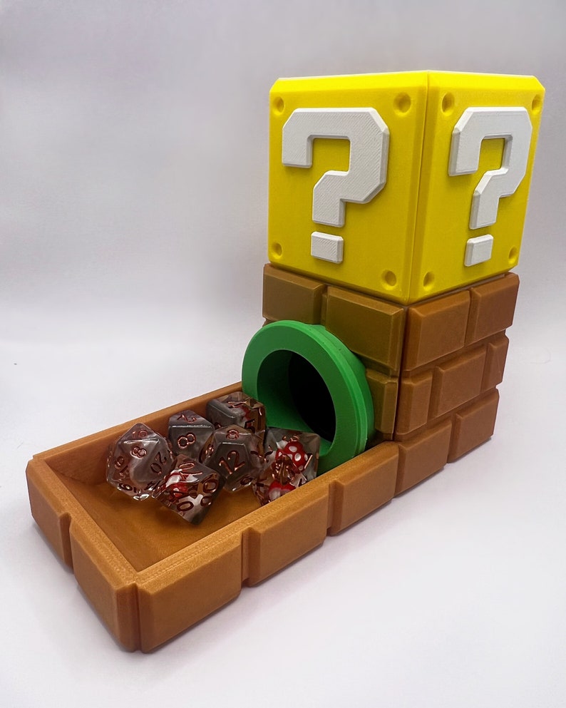 Op de afbeelding: Een 3D-geprinte dobbelsteentoren met bak, bestaande uit een geel vraagtekenblok op een bruine bakstenen basis met een groene buisopening. Verschillende doorschijnende rood-witte veelzijdige dobbelstenen met gouden cijfers liggen in de bak.