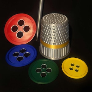 May include: A silver thimble with a gold band sits on a black surface. Four buttons are arranged around the thimble, one red, one blue, one green, and one yellow.
