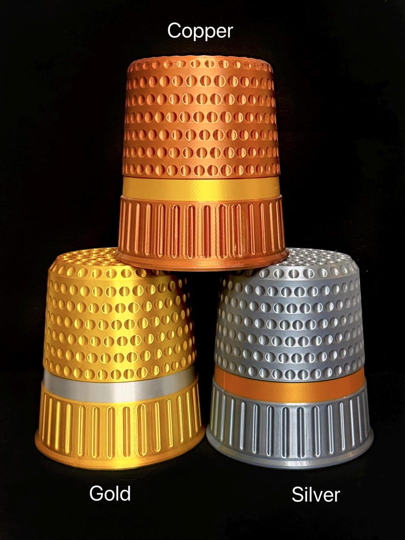 Op de afbeelding: Drie metalen vingerhoeden in koper, goud en zilver. De koperen vingerhoed staat bovenaan, met een gouden band. De gouden vingerhoed staat eronder, met een zilveren band. De zilveren vingerhoed staat rechts, met een oranje band. De woorden "Koper", "Goud" en "Zilver" staan op de afbeelding.