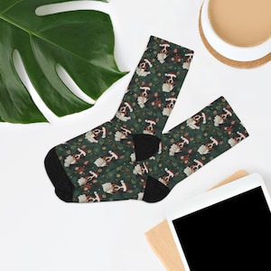 May include: A pair of dark green socks with a repeating pattern of dogs wearing Santa hats and festive greenery. The socks have black heels and toes. The socks are displayed on a white surface with a green leaf, a cup, and a tablet.