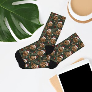 May include: A pair of Christmas-themed socks featuring a repeating pattern of a dog wearing a Santa hat, red ornaments, and sprigs of evergreen on a dark green background. The socks have black heels and toes.