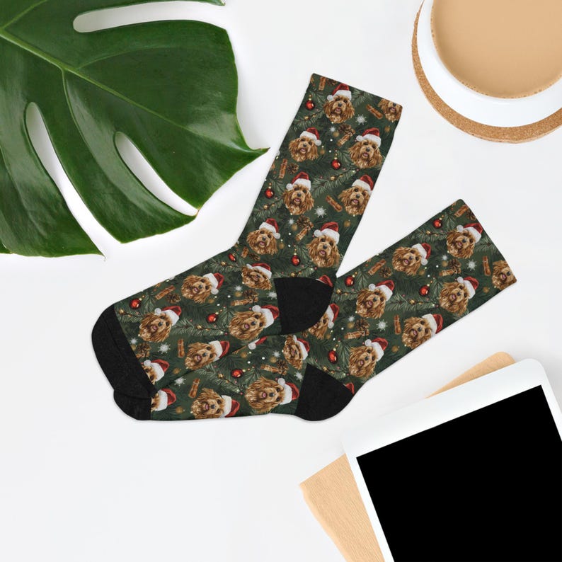 K&ouml;nnte beinhalten: Ein Paar Weihnachtssocken mit einem sich wiederholenden Muster eines Hundes mit Weihnachtsm&uuml;tze, roten Kugeln und Tannenzweigen auf dunkelgr&uuml;nem Hintergrund. Die Socken haben schwarze Fersen und Zehen.