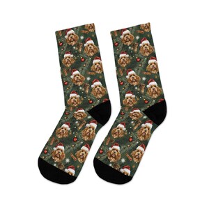 K&ouml;nnte beinhalten: Ein Paar Socken mit Weihnachtsmotiv, die ein sich wiederholendes Muster eines Hundes mit Weihnachtsm&uuml;tze, roten Ornamenten und Tannennadeln auf dunklem Gr&uuml;n zeigen. Die Socken haben schwarze Fersen und Zehen.