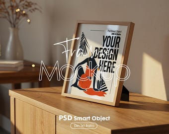 May include: A framed art print mockup with a wooden frame, displaying the text "YOUR DESIGN HERE." The artwork features a black and white illustration with orange and red accents. The frame is propped up on a wooden surface.