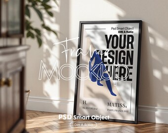 May include: A black framed art print mockup leaning against a wall. The print displays the text "YOUR DESIGN HERE", a blue abstract figure, and the text "MATISS" and "PARIS, FRANCE 1955". The frame is rectangular.