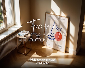 May include: A framed art print leans against a wall in a sunlit room. The artwork features a blue line drawing of a face with a red circle. A small side table holds magazines. The text "Frame Mockup" is overlaid on the image.