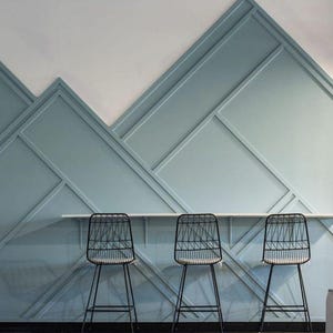 May include: Three black metal chairs with a geometric wire design sit in front of a light blue wall with a geometric pattern. The wall is made of wood panels.
