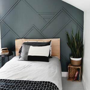 May include: A bedroom with a single bed with a wooden headboard and a gray and white patterned blanket. The wall behind the bed is painted a dark teal color and has a geometric pattern of wood trim. A potted plant sits on a small wicker table next to the bed.