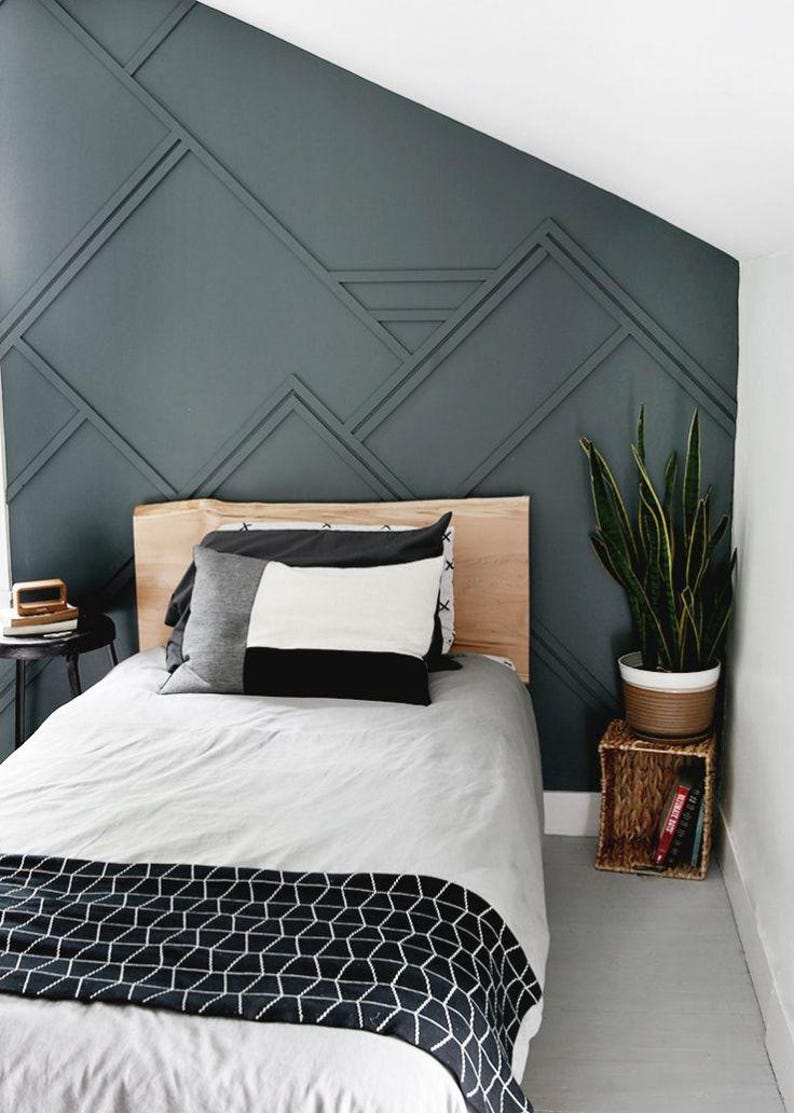 May include: A bedroom with a single bed with a wooden headboard and a gray and white patterned blanket. The wall behind the bed is painted a dark teal color and has a geometric pattern of wood trim. A potted plant sits on a small wicker table next to the bed.