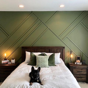 May include: A bedroom with a green accent wall featuring a geometric pattern. The wall is behind a bed with a white duvet and green and white pillows. A small black dog is lying on the bed. There are two nightstands with lamps on either side of the bed.