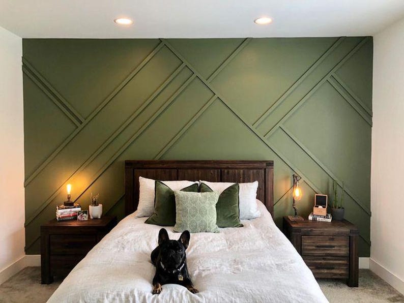 May include: A bedroom with a green accent wall featuring a geometric pattern. The wall is behind a bed with a white duvet and green and white pillows. A small black dog is lying on the bed. There are two nightstands with lamps on either side of the bed.