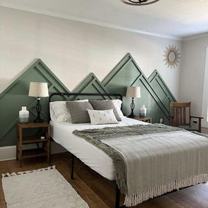 May include: A bedroom with a green and white mountain-shaped headboard behind a bed with a white duvet and a green and white throw blanket. There are two bedside tables with lamps and a chair in the corner.