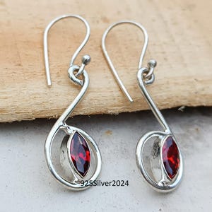 May include: A pair of silver earrings with a curved design. Each earring features a marquise-cut, deep red gemstone set within a silver frame. The earrings have a hook closure and are displayed on a wooden surface.