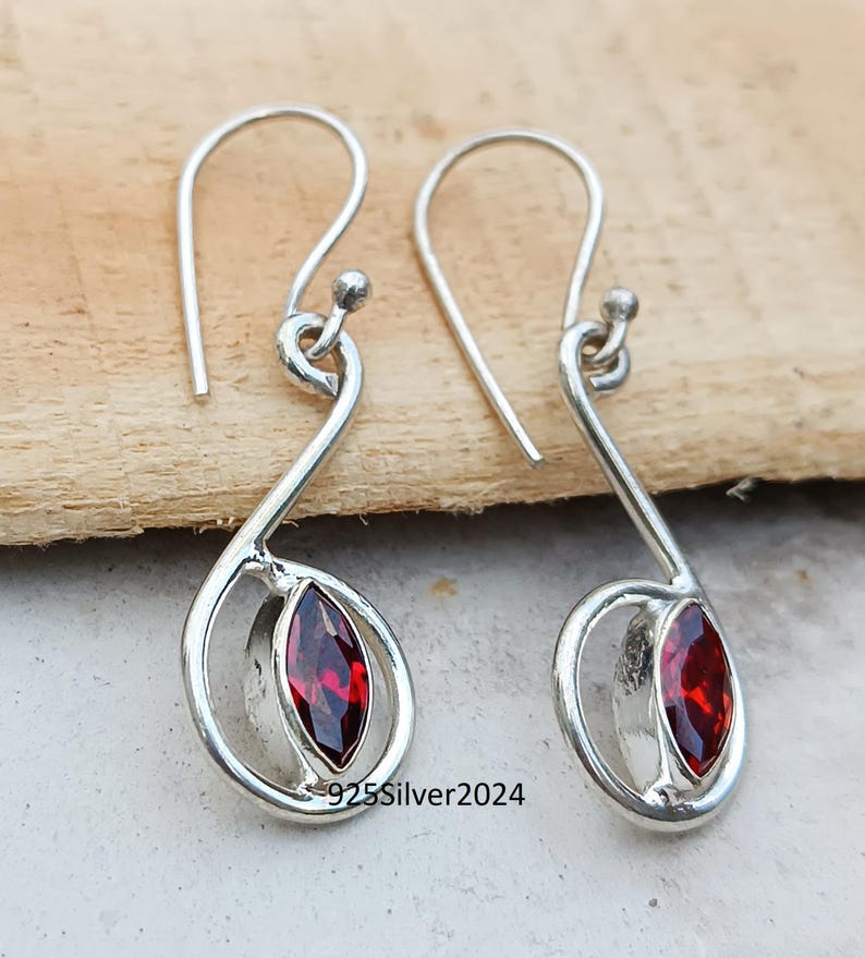 May include: A pair of silver earrings with a curved design. Each earring features a marquise-cut, deep red gemstone set within a silver frame. The earrings have a hook closure and are displayed on a wooden surface.