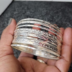Puede incluir: Un conjunto de siete pulseras de plata con diferentes diseños en relieve. Las pulseras están apiladas una encima de la otra y se sostienen en una mano.