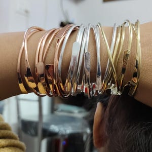 Puede incluir: Una pila de brazaletes de oro, plata y oro rosa. Los brazaletes tienen diferentes anchos y un acabado martillado.