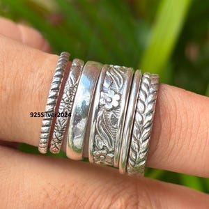 May include: A collection of silver rings with various designs, including floral, textured, and plain bands. The rings are stacked on a finger, showcasing the different styles and textures. The text "925Silver 2024" is visible.