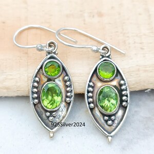 May include: A pair of silver earrings with a peridot gemstone design. Each earring features two green peridot stones, one oval and one round, set in a silver frame. The earrings have a hook closure and the text "925Silver2024".