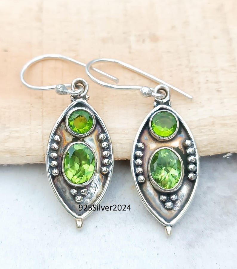 May include: A pair of silver earrings with a peridot gemstone design. Each earring features two green peridot stones, one oval and one round, set in a silver frame. The earrings have a hook closure and the text "925Silver2024".