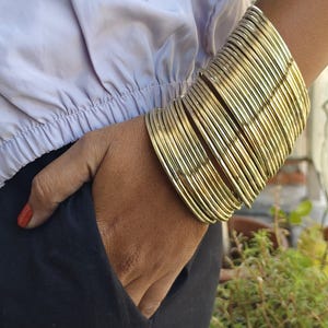 Puede incluir: Una pila de brazaletes de color dorado que se llevan en la muñeca de una persona. Los brazaletes están apilados en alto y crean un aspecto grueso y robusto.