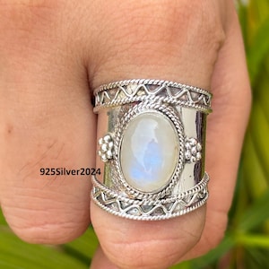 May include: A wide silver ring with an oval, iridescent moonstone center. The ring has decorative rope and geometric patterns. The text "925Silver2024" is visible on the side of the ring.