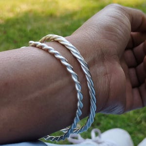 Può includere: Due braccialetti argentati, attorcigliati a forma di corda, sono indossati su un polso. I braccialetti sono in metallo lucido e hanno un aspetto intrecciato e strutturato. Lo sfondo è sfocato, con erba verde e un accenno di scarpa bianca visibile.