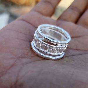 Może przedstawiać: Zestaw trzech srebrnych pierścionków ułożonych jeden na drugim. Górny pierścionek ma fakturę młotkową i rząd małych wycięć w kształcie serca. Środkowy pierścionek jest gładki, a dolny jest szerszy i ma lekko zaokrąglony brzeg.