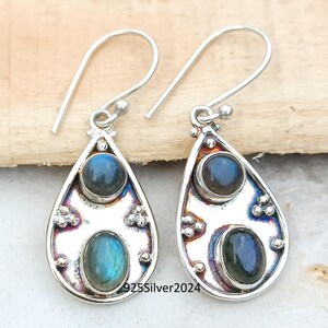 May include: A pair of silver teardrop earrings with two oval-shaped, iridescent blue gemstones. The earrings have a decorative border and a simple hook closure. The text "925Silver2024" is visible on the earrings.
