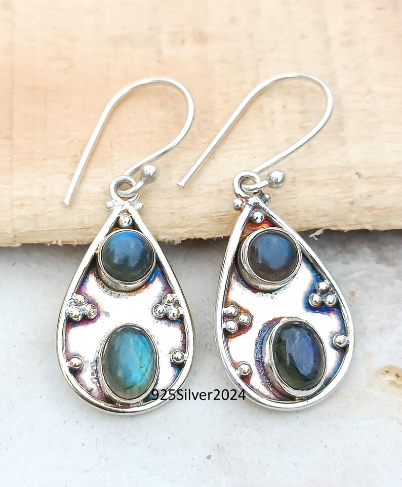 May include: A pair of silver teardrop earrings with two oval-shaped, iridescent blue gemstones. The earrings have a decorative border and a simple hook closure. The text "925Silver2024" is visible on the earrings.