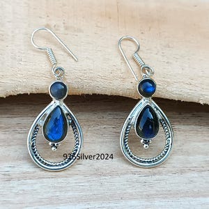 K&ouml;nnte beinhalten: Silberne Ohrringe mit blauen Edelsteinen. Jeder Ohrring hat einen kleinen, runden blauen Stein oben und einen gr&ouml;&szlig;eren, birnenf&ouml;rmigen blauen Stein in einem dekorativen Silberrahmen. Die Ohrringe haben einen Hakenverschluss. Der Text "925Silver2024" ist sichtbar.