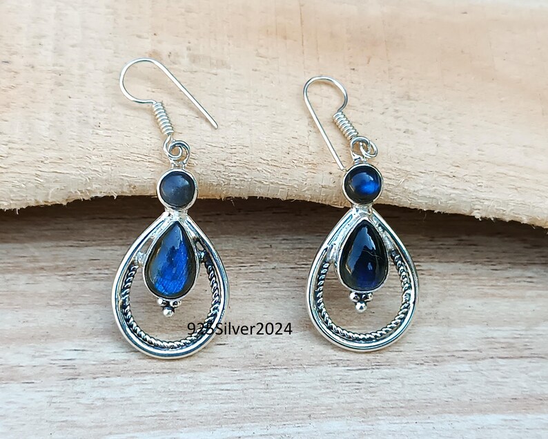 K&ouml;nnte beinhalten: Silberne Ohrringe mit blauen Edelsteinen. Jeder Ohrring hat einen kleinen, runden blauen Stein oben und einen gr&ouml;&szlig;eren, birnenf&ouml;rmigen blauen Stein in einem dekorativen Silberrahmen. Die Ohrringe haben einen Hakenverschluss. Der Text "925Silver2024" ist sichtbar.