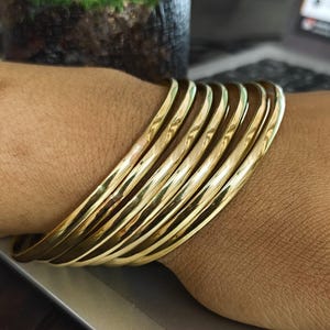 Op de afbeelding: Een stapel goudkleurige armbanden om een pols. De armbanden zijn glad en glanzend en reflecteren licht. De armbanden zijn op elkaar gestapeld, waardoor een gelaagd effect ontstaat. De sieraden hebben een warme goudkleur.