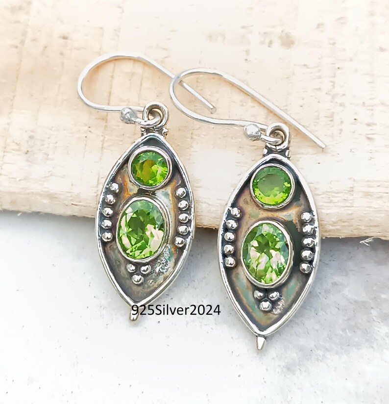 May include: A pair of sterling silver earrings with a drop design. Each earring features two oval-cut, light green gemstones. The earrings have a hook closure and are embellished with small silver beads. The text "925Silver2024" is visible.