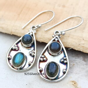May include: A pair of silver teardrop earrings with two oval-shaped, dark blue and iridescent blue-green gemstones. The earrings have a decorative design around the stones and a hook closure. The text "925Silver2024" is visible.
