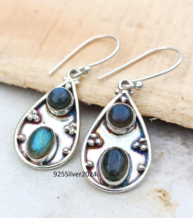 May include: A pair of silver teardrop earrings with two oval-shaped, dark blue and iridescent blue-green gemstones. The earrings have a decorative design around the stones and a hook closure. The text "925Silver2024" is visible.