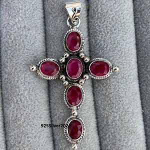 Può includere: Un ciondolo a forma di croce in argento, ornato da pietre preziose ovali di colore rosso intenso. Le pietre sono incastonate in un bordo dettagliato a forma di corda. Il ciondolo ha un anello nella parte superiore per una catena. Il testo "925Silver202" è visibile.