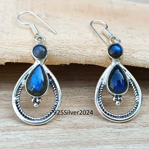 K&ouml;nnte beinhalten: Silberne Ohrringe mit tropfenf&ouml;rmigen blauen Edelsteinen. Die Ohrringe haben einen kreisf&ouml;rmigen oberen Teil und ein dekoratives, gedrehtes Silberdesign um den Edelstein. Der Text "925Silver2024" ist auf der Holzoberfl&auml;che sichtbar.