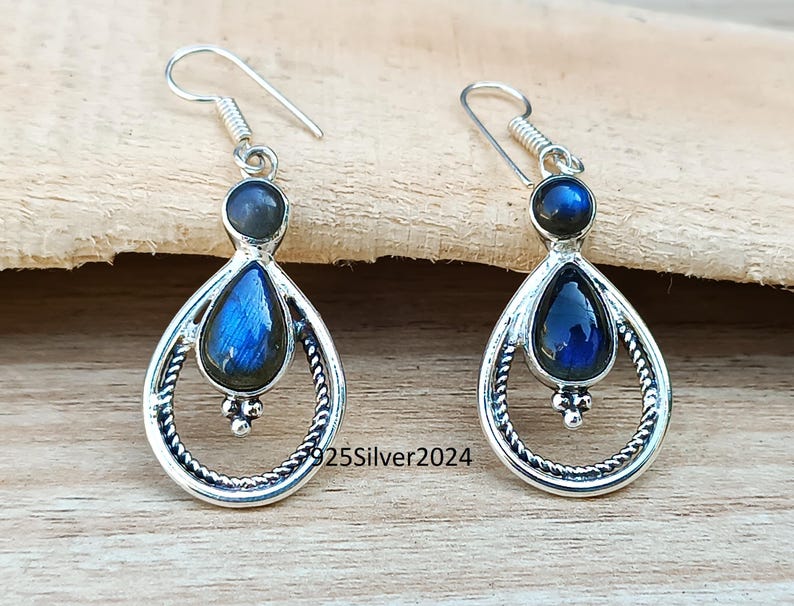 K&ouml;nnte beinhalten: Silberne Ohrringe mit tropfenf&ouml;rmigen blauen Edelsteinen. Die Ohrringe haben einen kreisf&ouml;rmigen oberen Teil und ein dekoratives, gedrehtes Silberdesign um den Edelstein. Der Text "925Silver2024" ist auf der Holzoberfl&auml;che sichtbar.