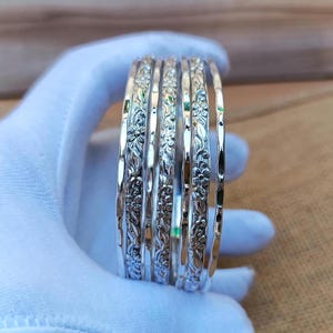 Pode incluir: Um conjunto de seis pulseiras de prata com padrões florais. As pulseiras são seguradas em uma mão enluvada, destacando seus intrincados desenhos. As pulseiras têm um acabamento polido e reflexivo, com delicadas gravuras florais.