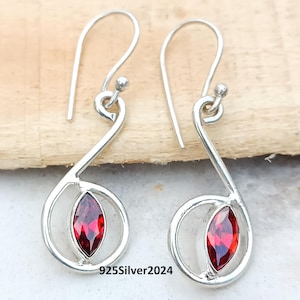 May include: A pair of silver earrings with a teardrop design. Each earring features a vibrant red gemstone set within a swirling silver frame. The earrings have a hook closure and are displayed on a wooden surface. The text "925Silver2024" is visible.