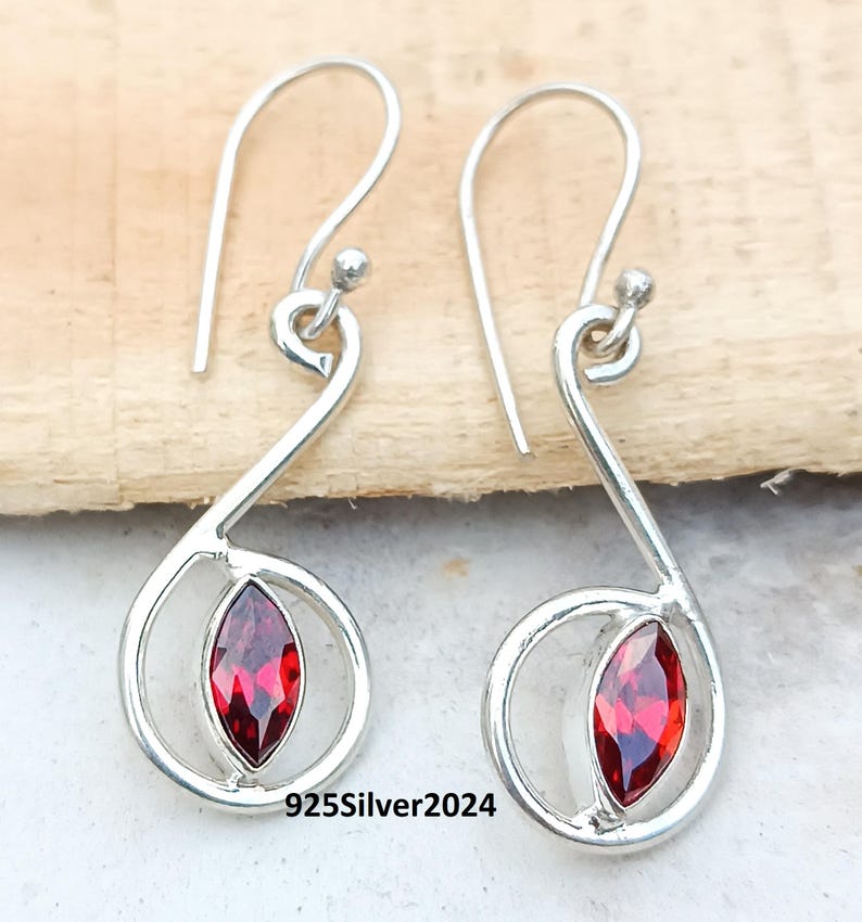 May include: A pair of silver earrings with a teardrop design. Each earring features a vibrant red gemstone set within a swirling silver frame. The earrings have a hook closure and are displayed on a wooden surface. The text "925Silver2024" is visible.