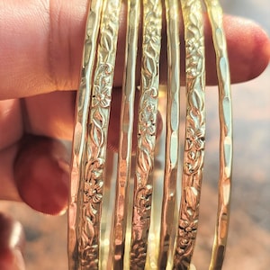 Puede incluir: Un conjunto de siete pulseras doradas con acabado martillado y diseños florales. Las pulseras están apiladas y son un accesorio popular para mujeres.