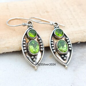 May include: A pair of sterling silver dangle earrings. Each earring features two green peridot gemstones set in an oval, textured silver frame. The earrings have a hook closure. The text "925Silver2024" is visible.