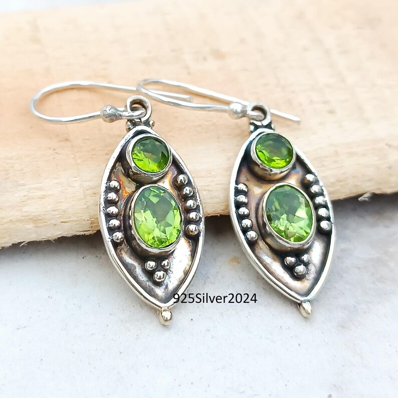 May include: A pair of sterling silver dangle earrings. Each earring features two green peridot gemstones set in an oval, textured silver frame. The earrings have a hook closure. The text "925Silver2024" is visible.