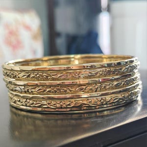 May include: Set of five gold-toned bangle bracelets with a floral design. The bracelets are stacked on top of each other.